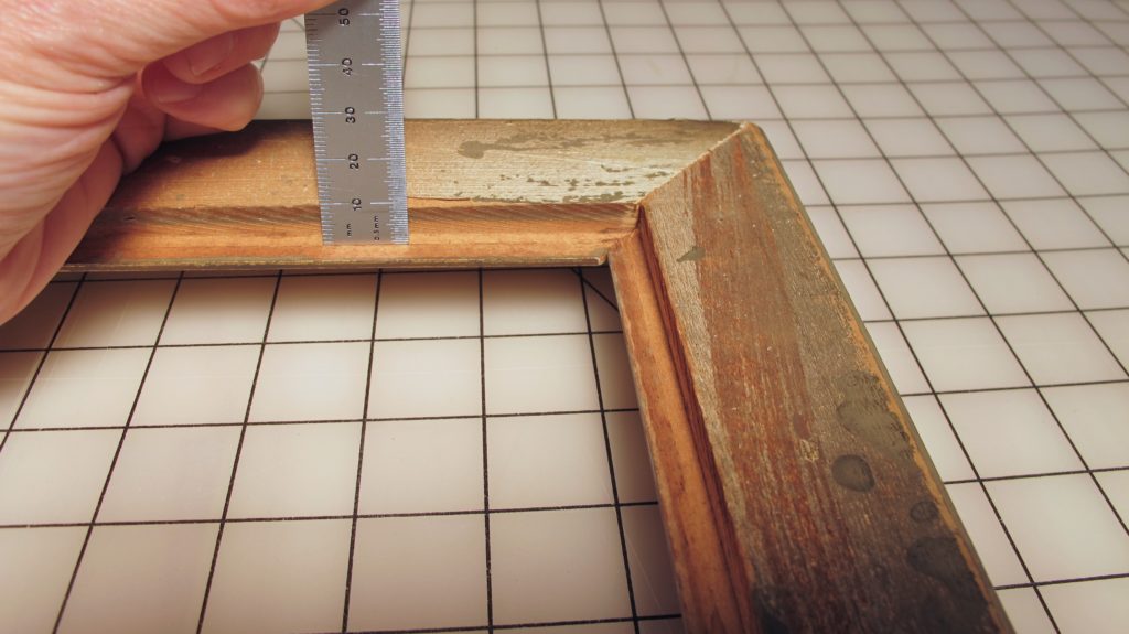 Hand measuring the depth of an aged wooden frame with a metal ruler.