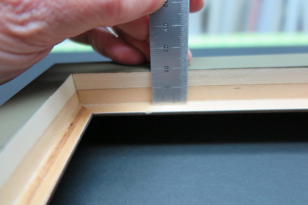 Hand using a metal ruler to measure the depth of a wooden frame.