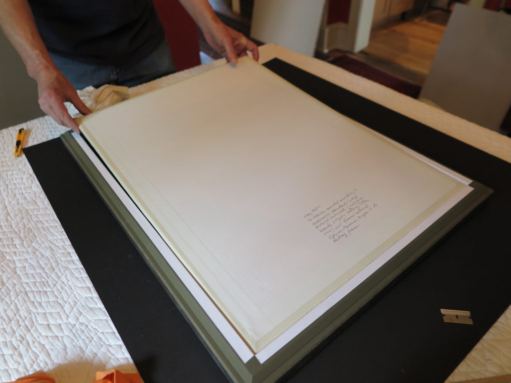 Hands placing a textile-mounted artwork package into a prepared frame.