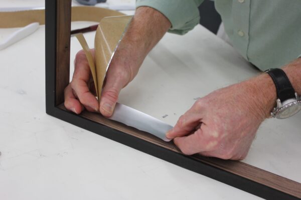 Hands applying Marvelseal barrier to the inside edge of a wooden frame for artwork protection.