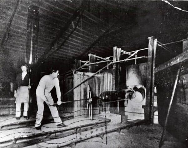 Historic glassmaking process showing artisans working with molten glass in an early industrial workshop.