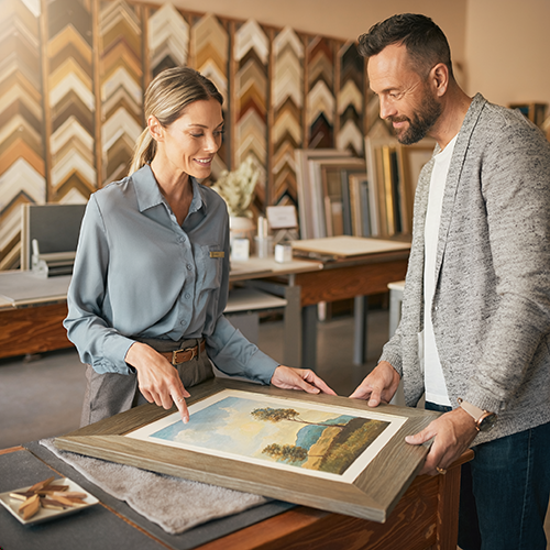 Professional framer pointing to UV-protective glass in a picture frame, explaining how custom framing helps preserve artwork and prevent fading.